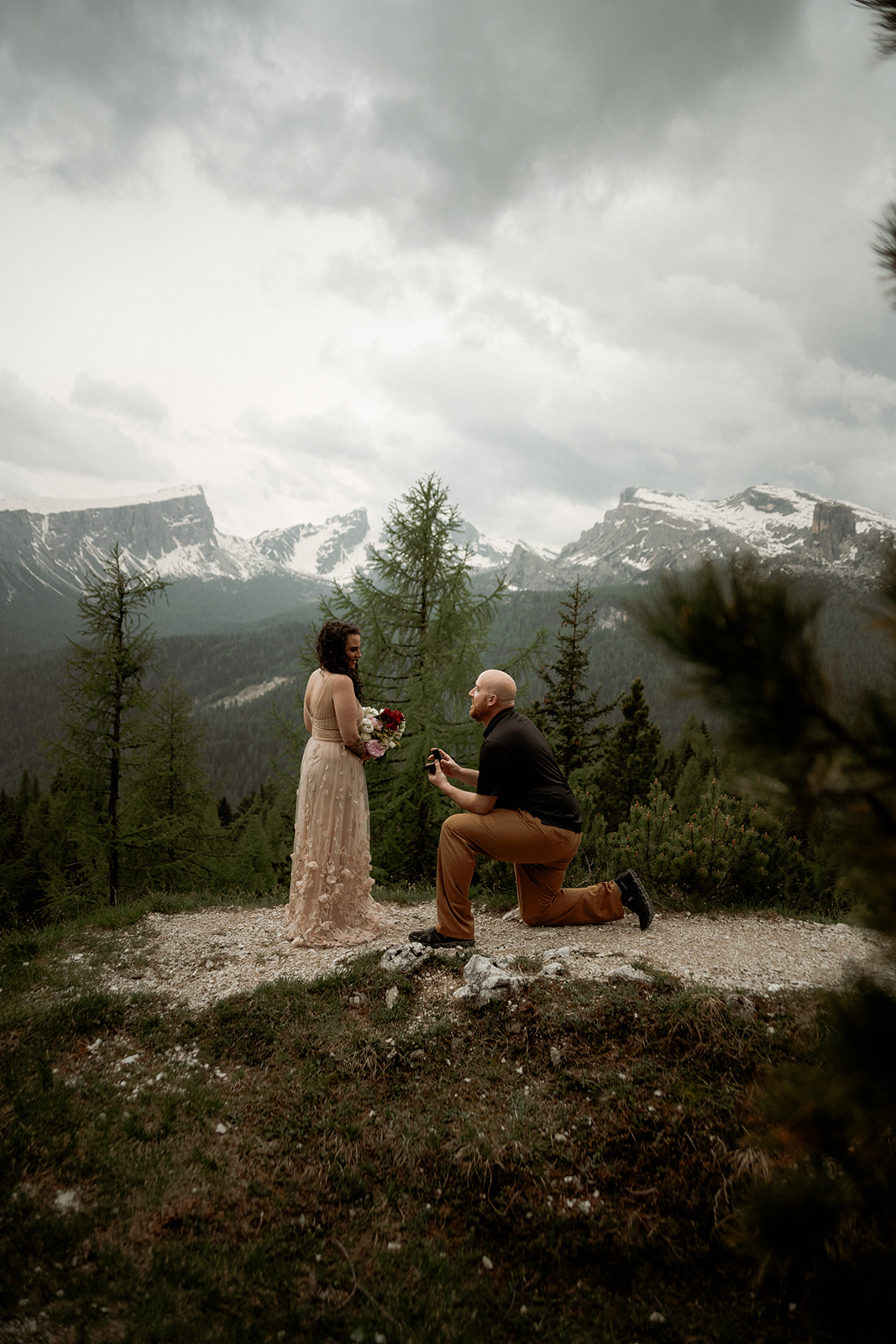Heiratsantrag in den Bergen in einer Waldlichtung mit dolomiten im Hintergrund, Frau hält geschenktem Blumenstrauß in der Hand