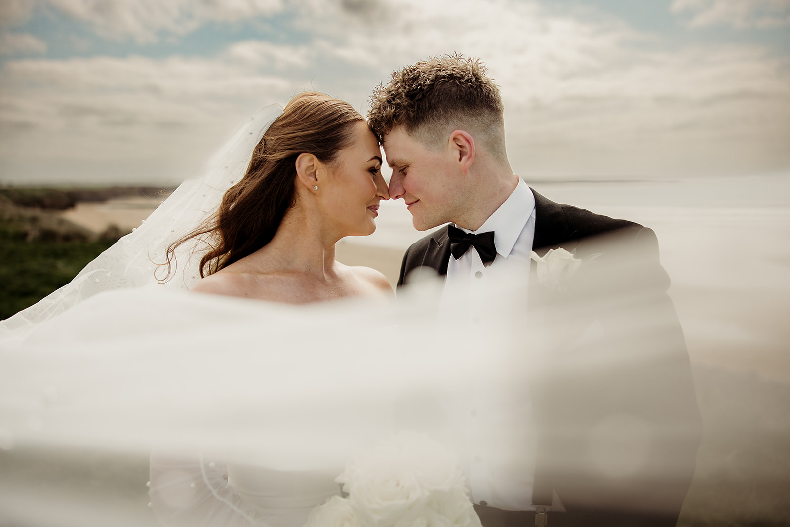 Alison and Ross together by the sea, with her veil blowing across the foreground