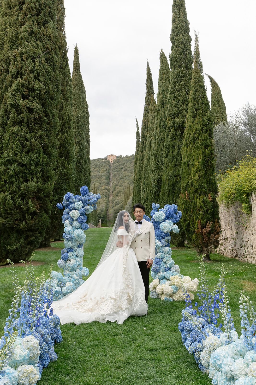 Bride and groom at a ceremony framed by blue hydrangeas and cypress trees