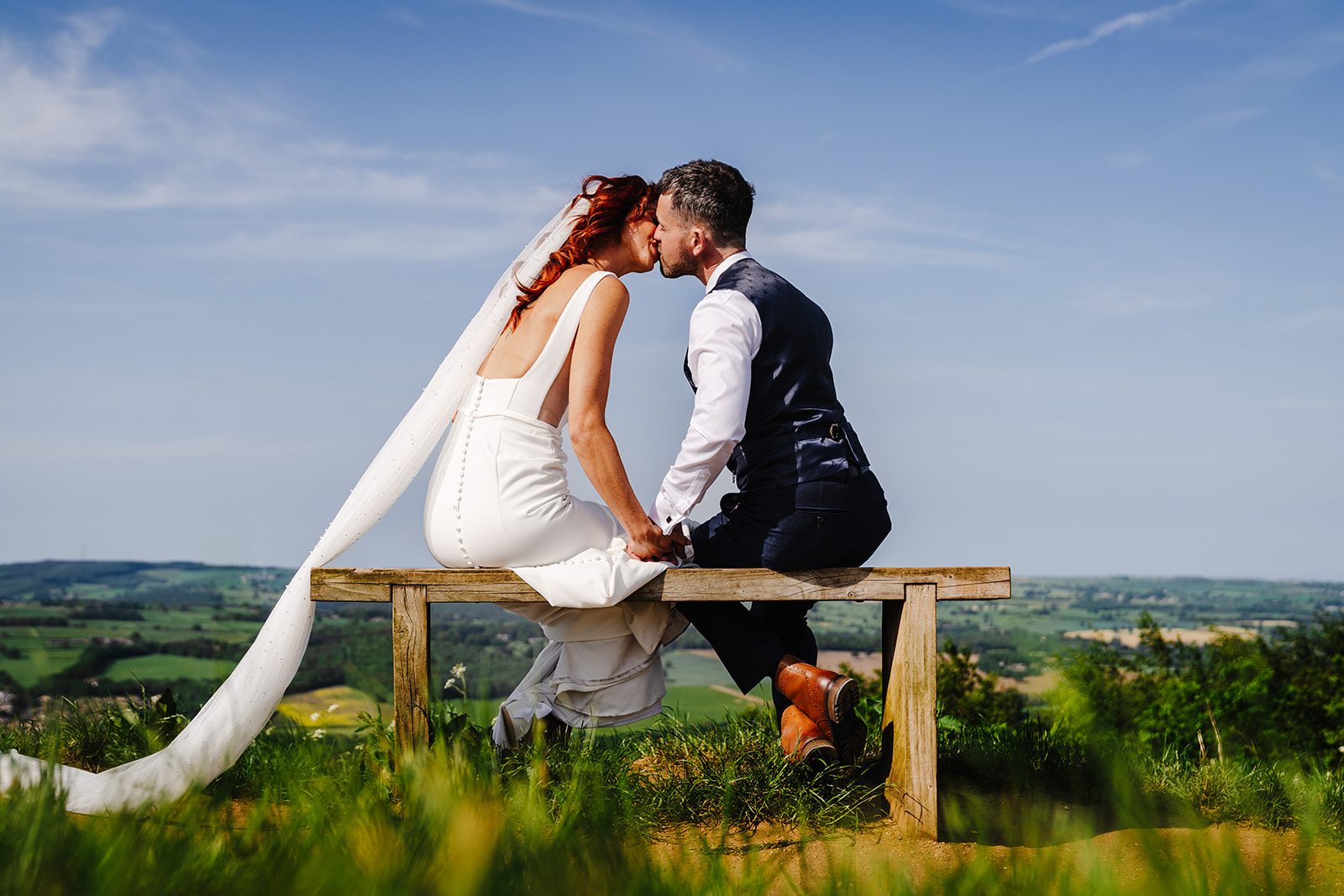Nicky and Luke kissing on a bench with countryside behind them