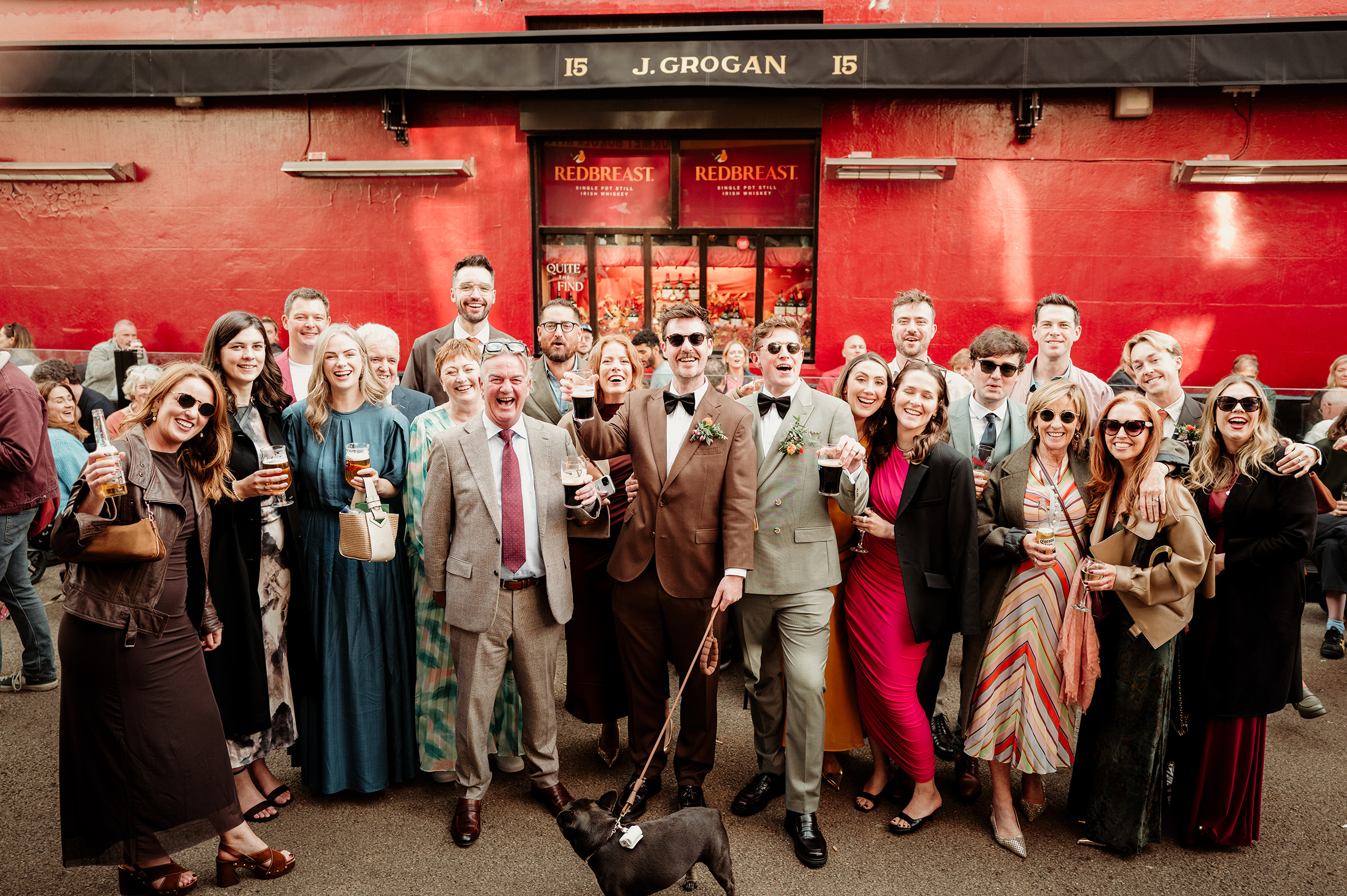 Two grooms celebrating outside a red Dublin pub with their wedding guests and dog.