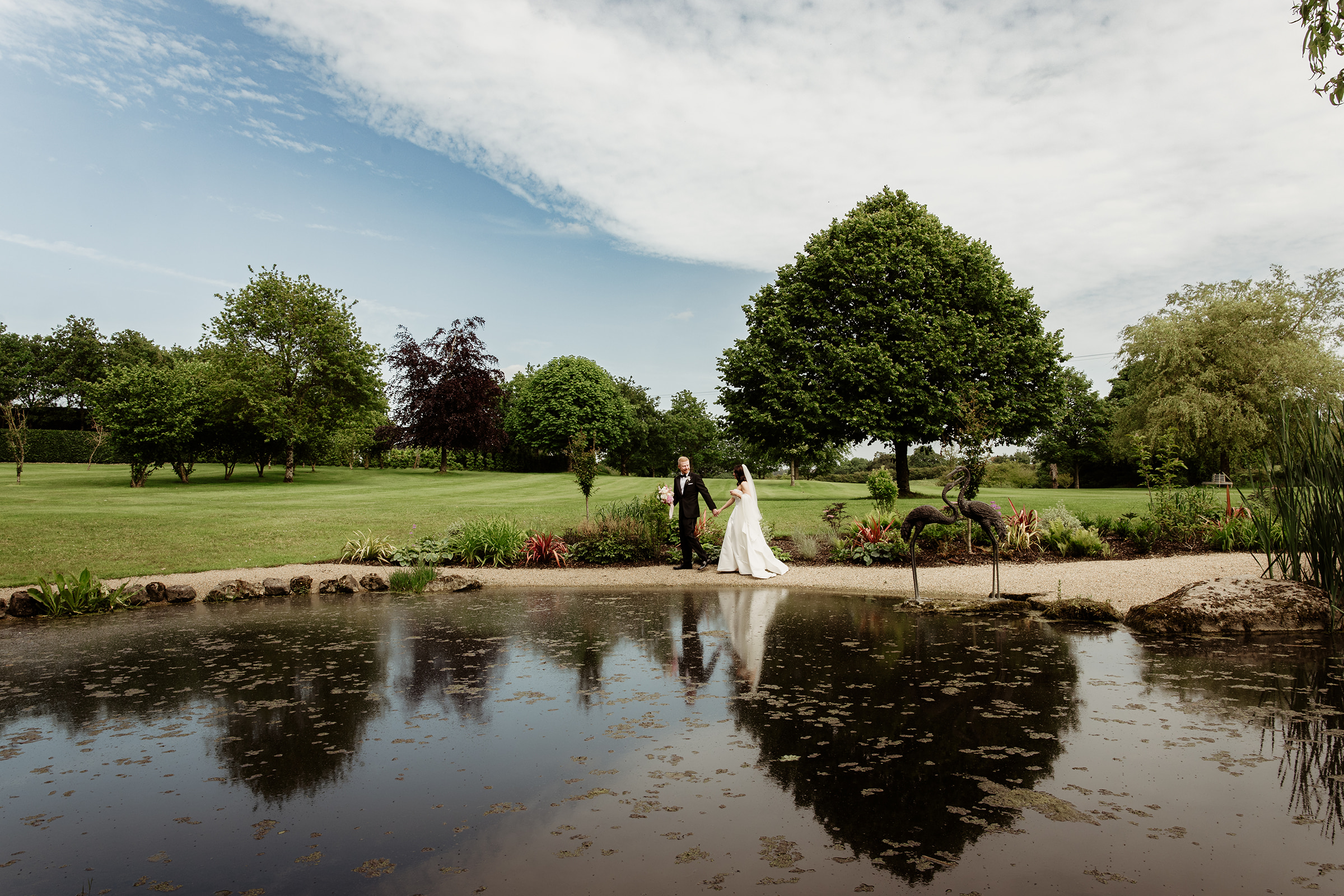 A newly married couple walking hand in hand beside a garden pond, reflected in the water.