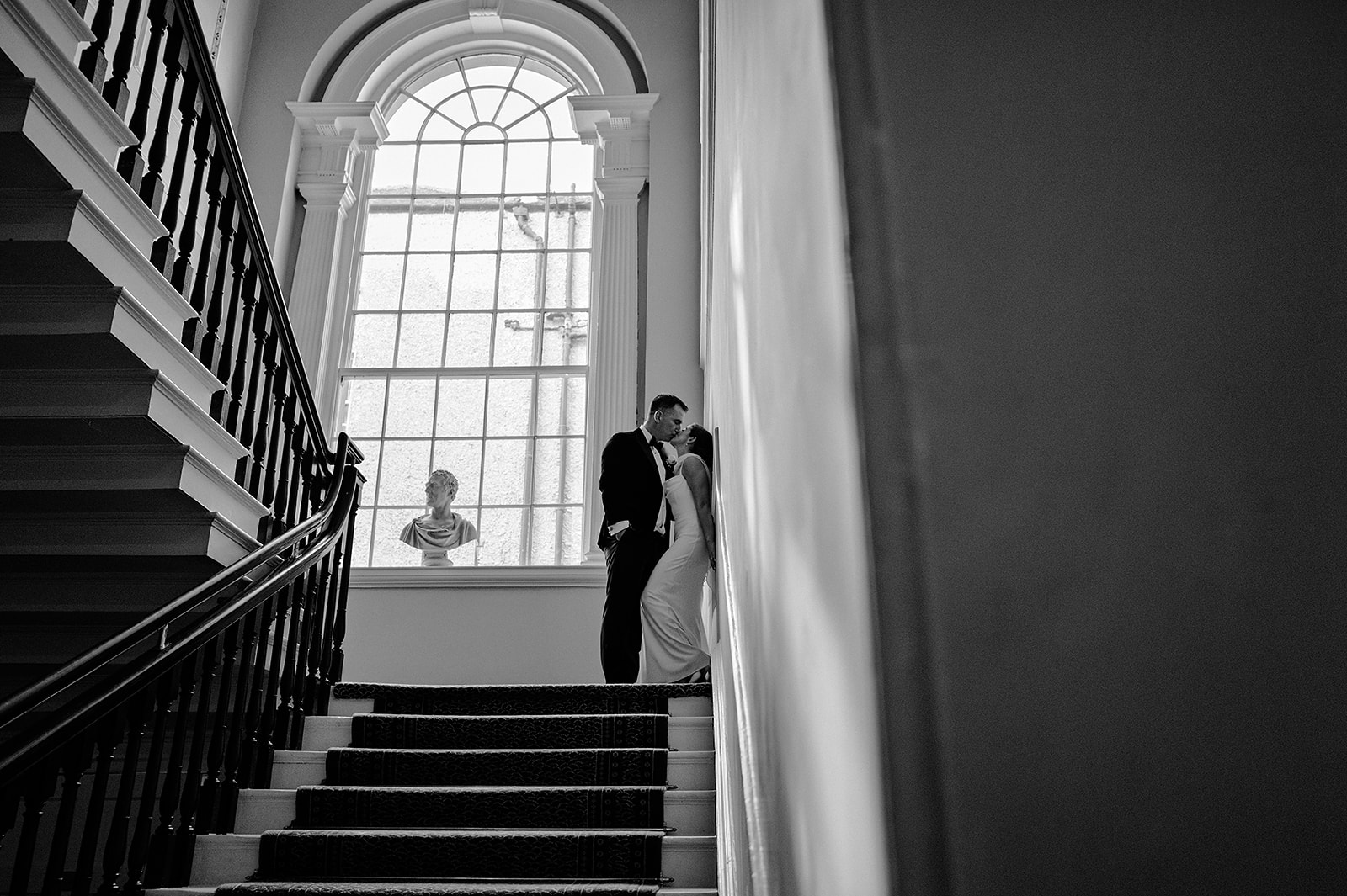 Rebecca and Louis kissing beside a tall arched window on a staircase, photographed in black and white